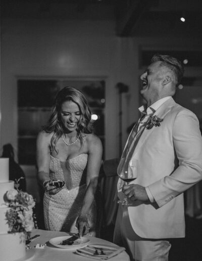 A couple laughs while cutting a cake at a wedding reception; the woman holds a piece of cake, and the man holds a glass.