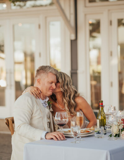 A couple sits at a table, embracing with closed eyes. A bottle of wine, wine glasses, and plates are on the table. The background features a building with large windows and doors.