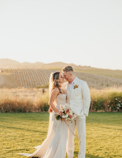 A couple dressed in wedding attire stands closely on a sunlit grassy field, with vineyards in the background.