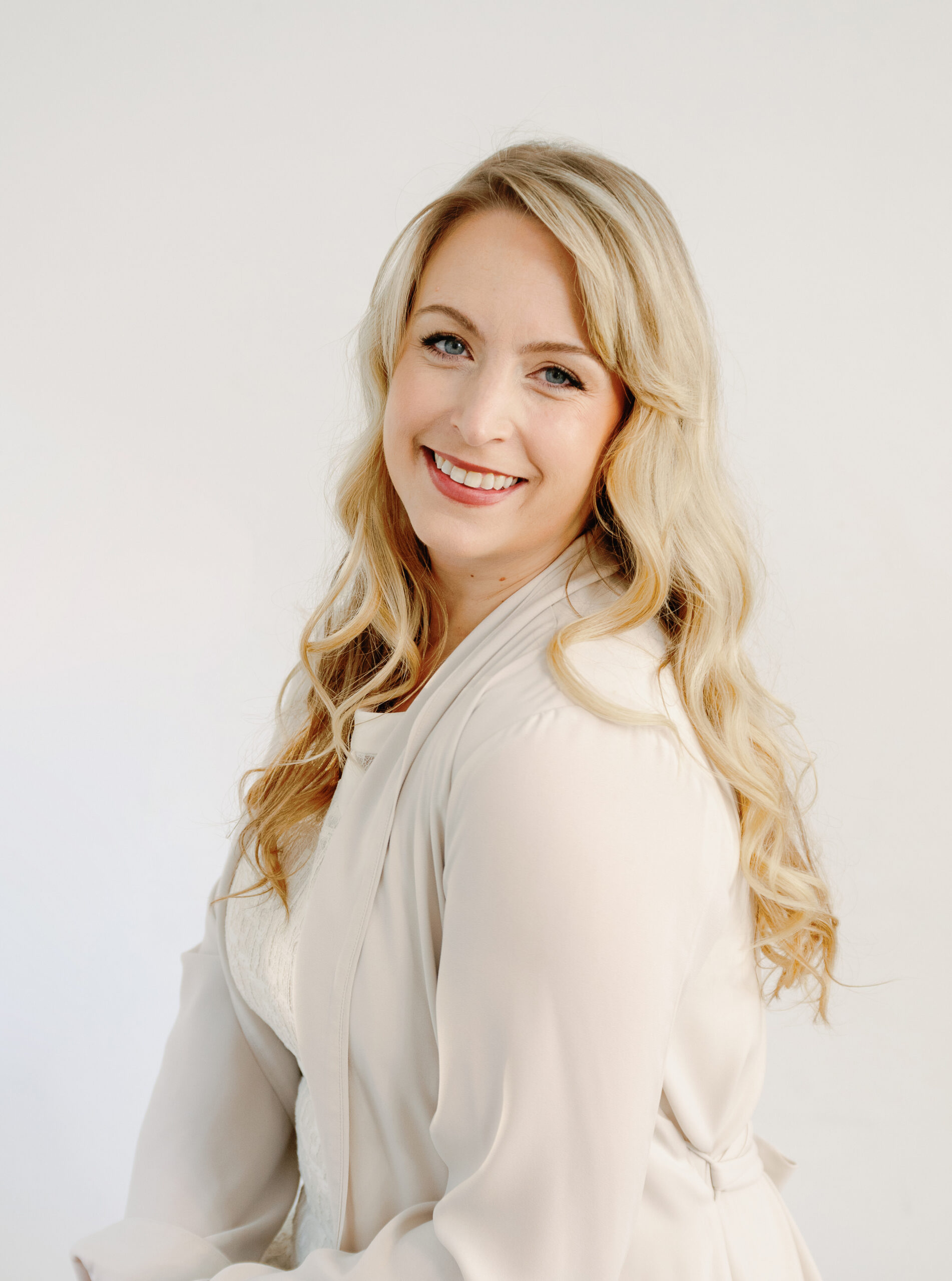 Woman with long blonde hair, wearing a light-colored blouse, smiling against a plain background.