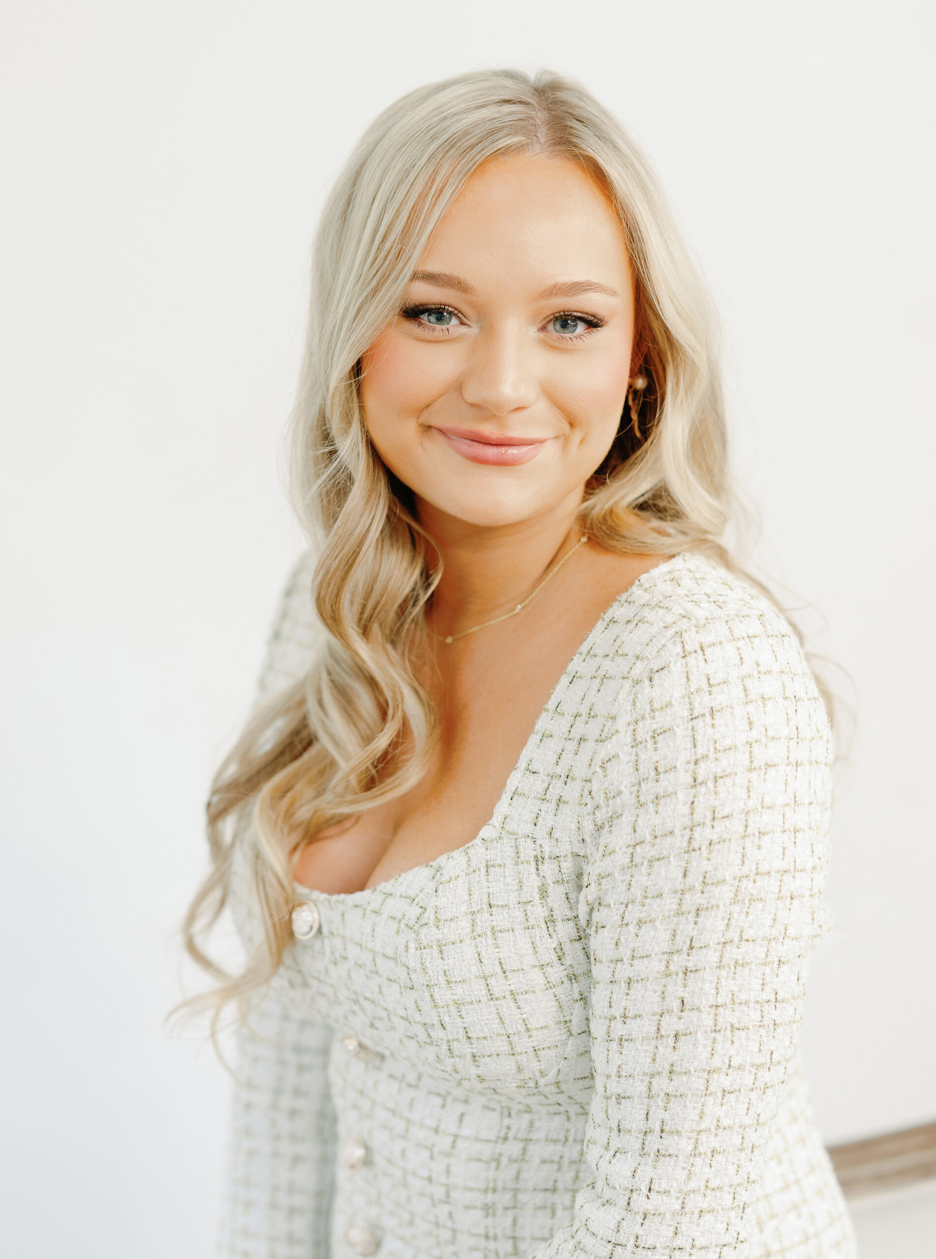 A woman with long blonde hair and a white textured top smiles softly against a plain background.