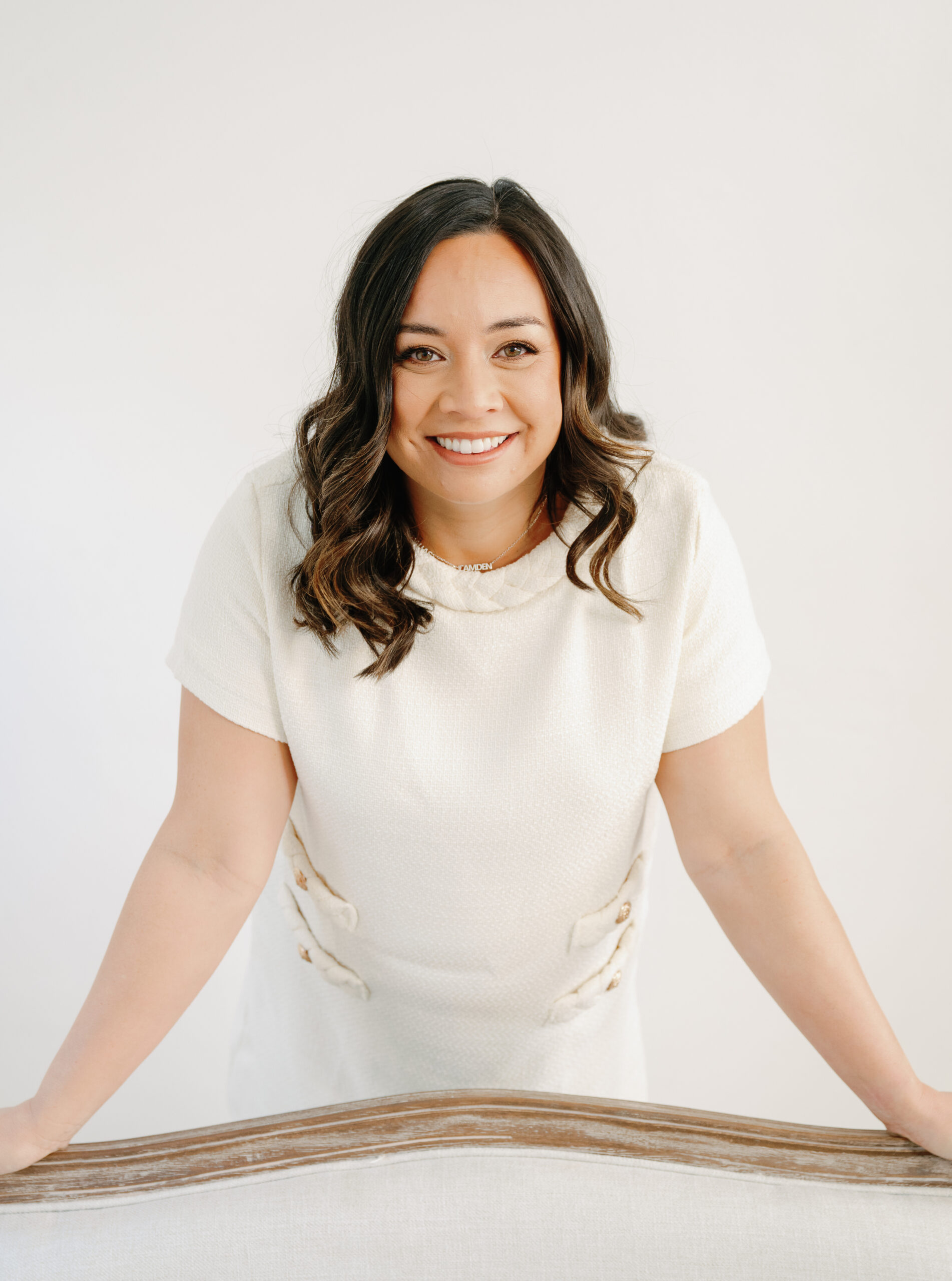 A woman in a white dress leans on a wooden railing, smiling at the camera against a neutral background.