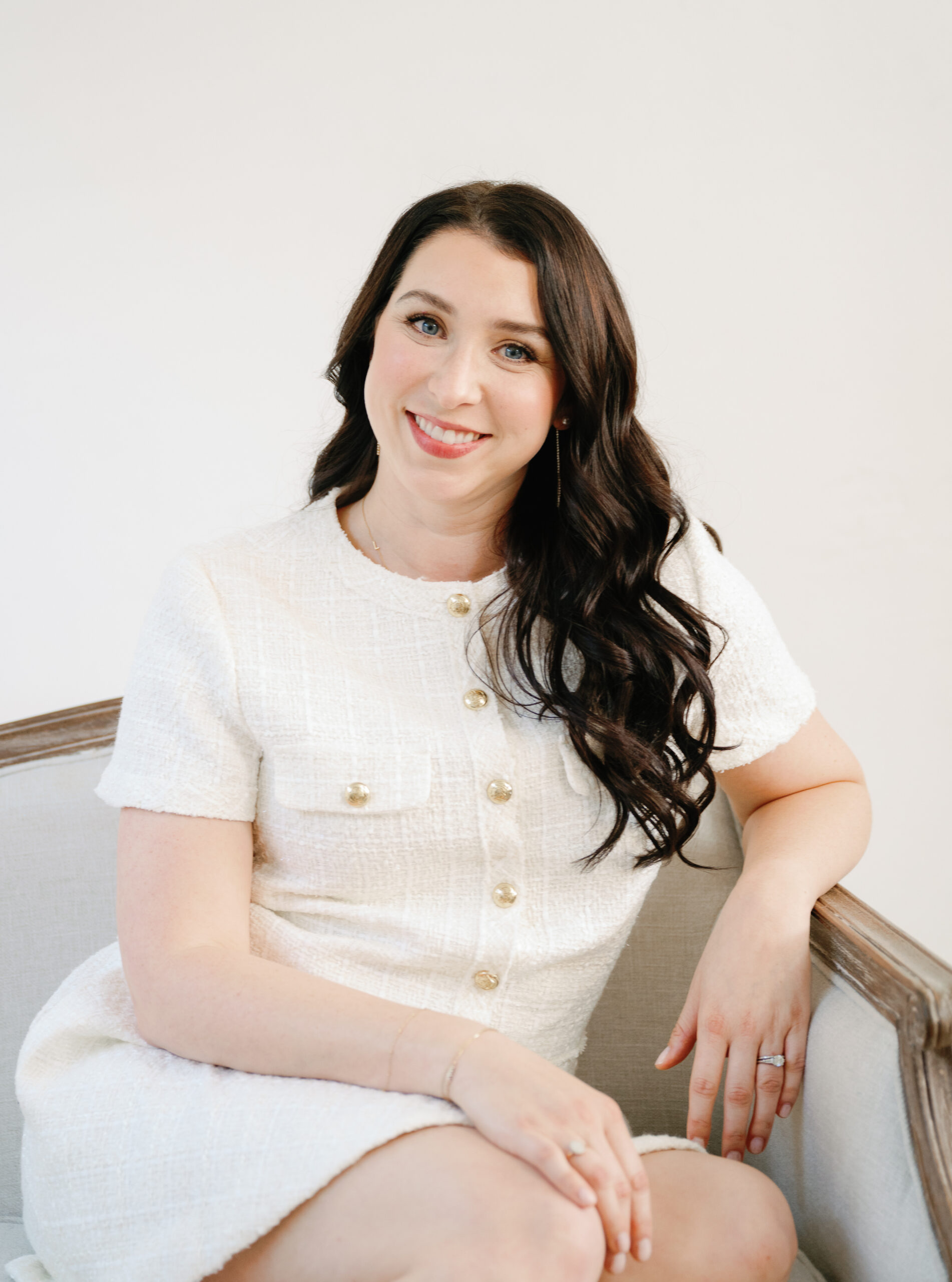 A woman with long dark hair sits in a light-colored chair, wearing a white textured short-sleeve dress. She is smiling and resting her arm on the armrest.