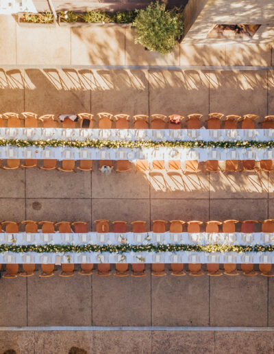 Aerial view of two long tables set up outdoors with white tablecloths and floral centerpieces, surrounded by wooden chairs.