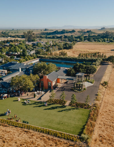 Aerial view of a rural property with buildings, a lawn area, and surrounding fields. People are visible on the lawn, and hills extend into the distance.