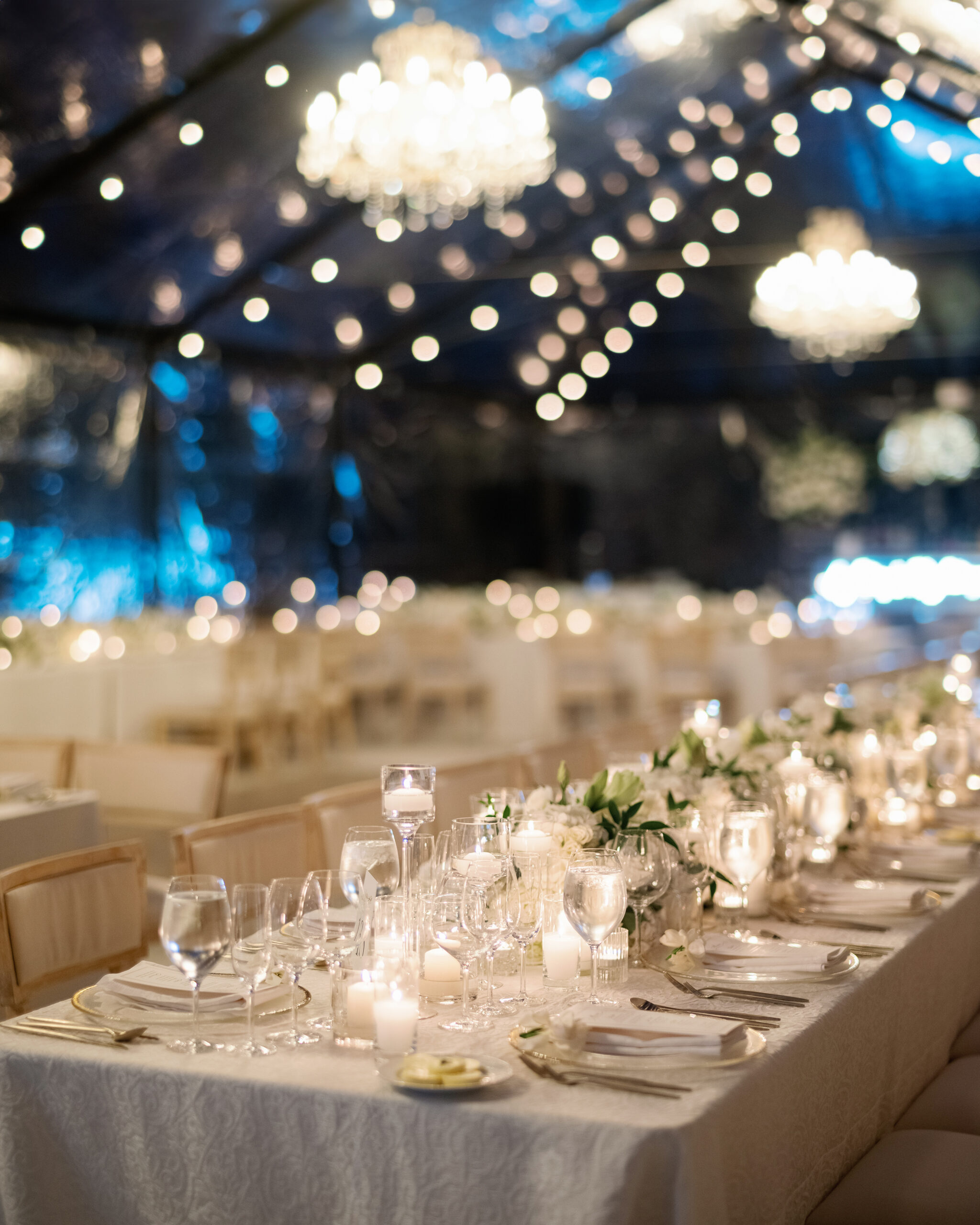 Elegant indoor dining setup with long tables, white tablecloths, floral centerpieces, glassware, and chandeliers overhead, creating a formal and ambient atmosphere.