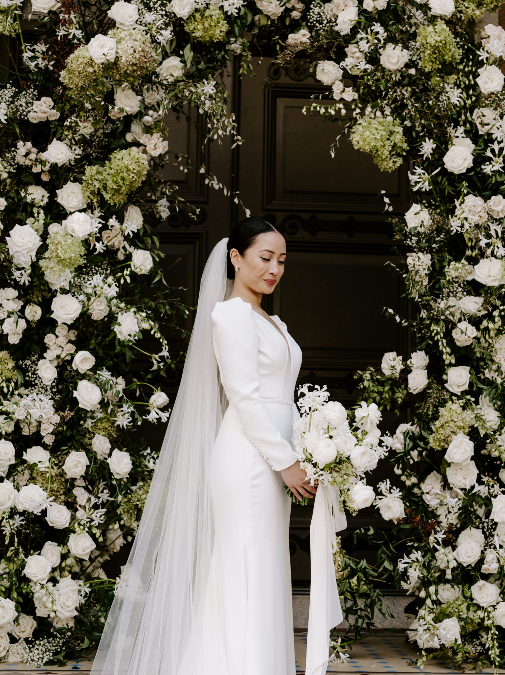 A bride stands in front of a floral arch, holding a bouquet. She wears a long-sleeved white dress and a veil.