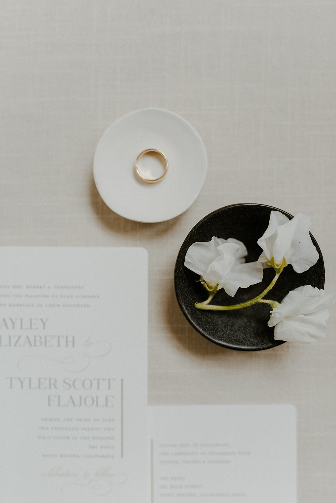 A flat lay image features a gold ring on a small dish, white flowers in a black bowl, and wedding invitations on a light tablecloth.