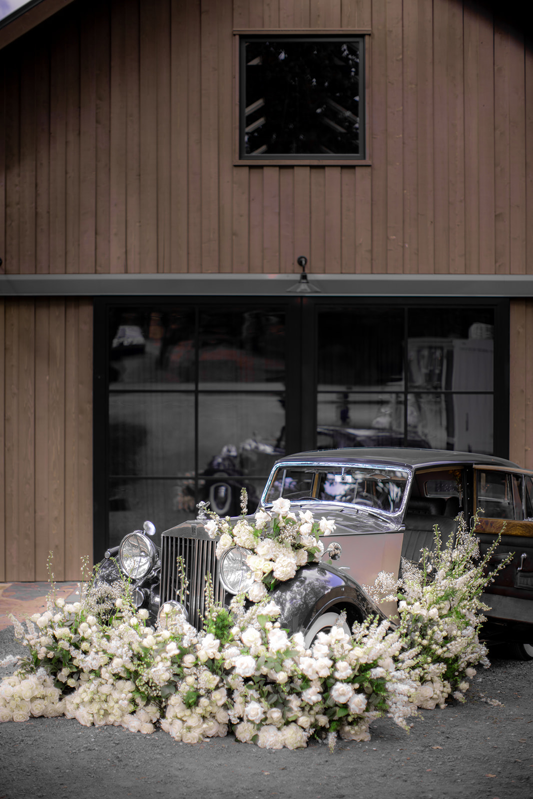 A vintage car is adorned with an abundance of white flowers in front of a wooden building with large glass doors.