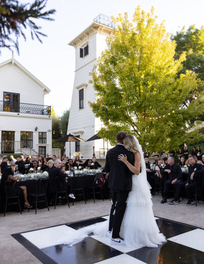 A couple dances on a black and white checkered floor outdoors, surrounded by seated wedding guests in front of a large white building and a yellow-leaved tree.