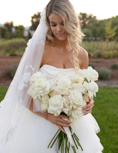 Bride in a white gown holding a bouquet of white roses, standing on grass with trees in the background.