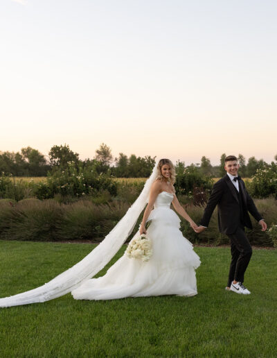 A bride and groom walk hand in hand on a grassy field. She wears a long white gown with a veil, holding a bouquet. He wears a black suit and sneakers. Trees line the background.