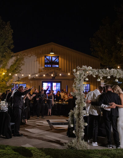 People gather outdoors at night in front of a barn-style building, celebrating under string lights. A small group embraces under a floral arch.