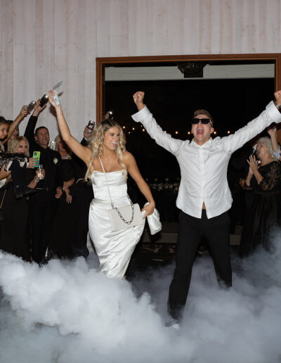 A couple celebrating energetically on a dance floor surrounded by smoke, with applauding guests in the background.