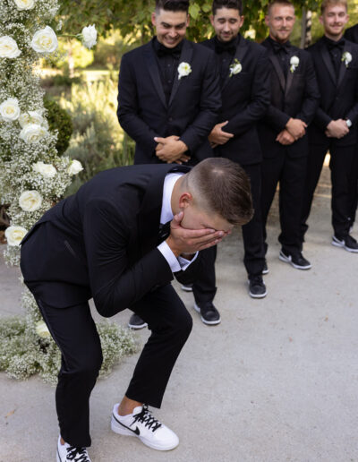 A person in a black suit and white sneakers covers their face, surrounded by four similarly dressed individuals and floral decorations.