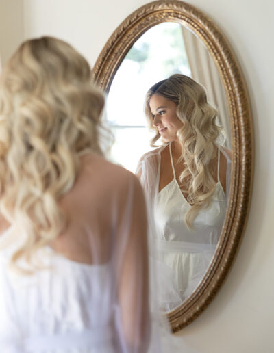 A woman in a white dress stands in front of an oval mirror, gazing at her reflection. Her wavy blonde hair falls over her shoulders.