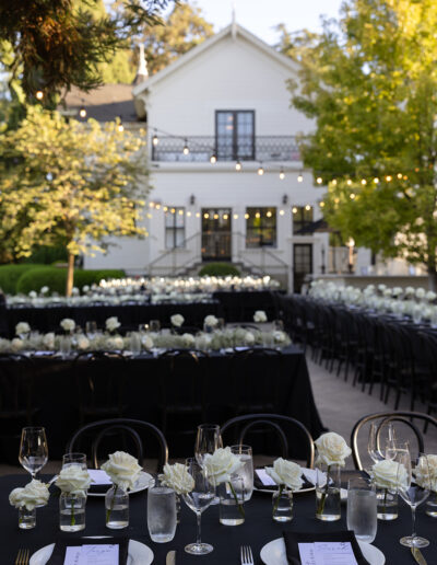 Outdoor wedding reception setup with black tablecloths and white roses on tables in front of a white house with string lights above.