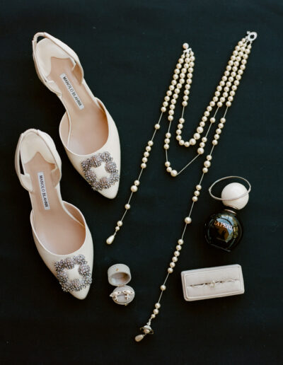 A pair of embellished white shoes, two pearl necklaces, a La Perla perfume bottle, a ring in a box, and a pen on a black surface.