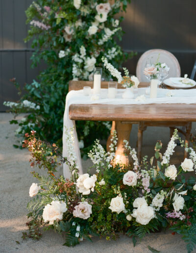 Elegant wedding table with floral arrangement, white candles, and draped fabric surrounded by greenery and pale roses.