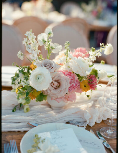 A floral centerpiece featuring pink, white, and yellow flowers in a vase on a table set with plates and menus.