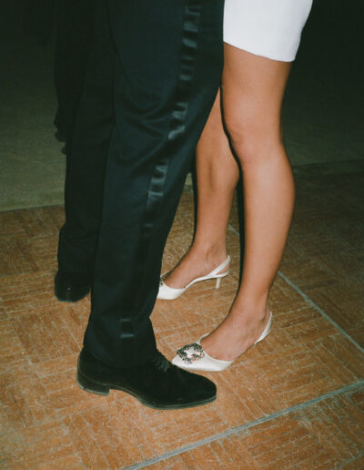 A couple dances closely on a tiled floor, with the man in dark pants and the woman in a white dress and white heels.