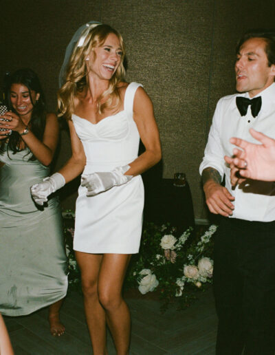 A group of people in formal attire dancing at an indoor event. A woman in a white dress and gloves is smiling in the center.