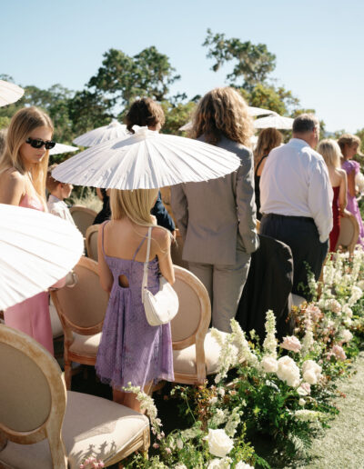 Wedding ceremony guests stand outdoors with parasols, surrounded by floral arrangements.