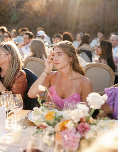 A woman in a pink dress sits at an outdoor event table, wiping her eye. The table is adorned with flowers and glasses. Other guests are seated around her.