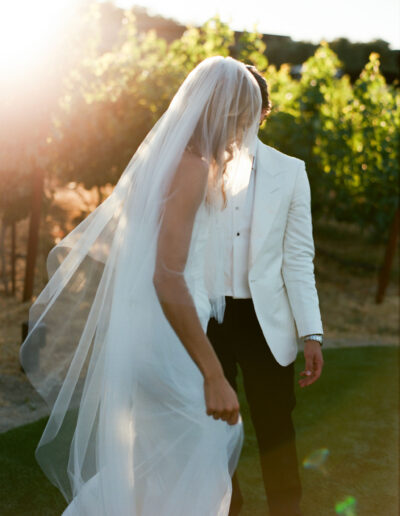 A bride and groom stand outdoors, with the bride in a long veil and white dress, and the groom in a white jacket. They are surrounded by sunlit greenery.