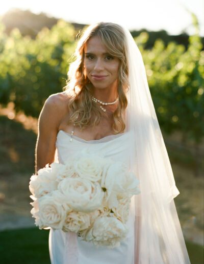 Bride in a white dress and veil holding a bouquet of white flowers, standing outdoors with greenery in the background.