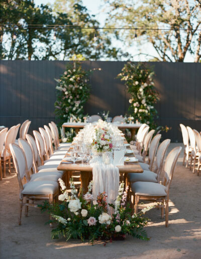 An elegantly set outdoor dining table with floral arrangements and white chairs under string lights.