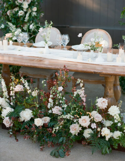 Elegant wooden table set for a formal event, adorned with white and blush flowers and candles, with three empty chairs.