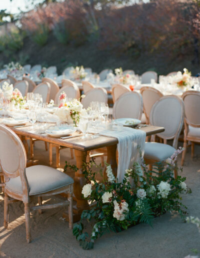 Outdoor event setting with wooden tables and beige upholstered chairs, adorned with white tablecloths, glassware, and floral centerpieces. Lush greenery surrounds the tables.