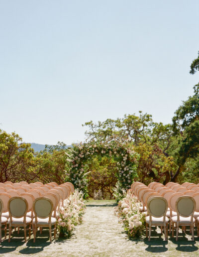 Outdoor wedding ceremony setup with rows of beige chairs facing a floral arch under a clear blue sky.