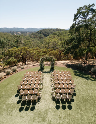 Outdoor wedding ceremony setup with rows of chairs on grass facing a floral arch, surrounded by lush greenery and hills in the background.