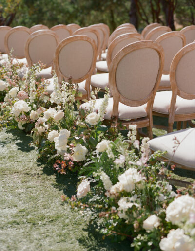 Rows of beige chairs are set up outdoors. The aisle is lined with white and pink flowers on green grass.