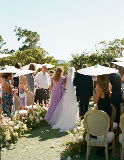 Bride walking down the aisle with a woman in a purple dress and a man in a suit, surrounded by wedding guests with parasols.