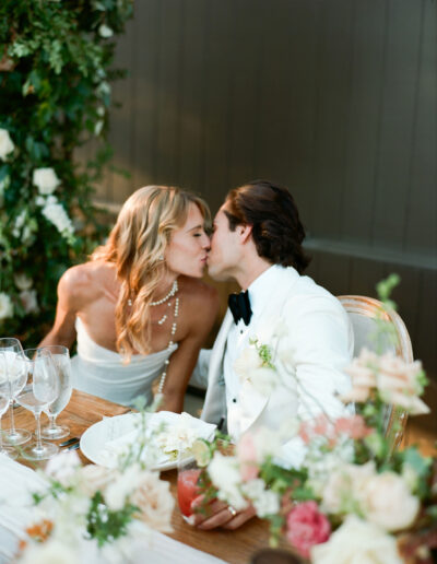 A couple in formal attire kisses at a flower-adorned table set with glassware and plates.