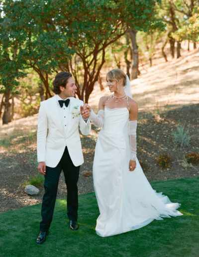 A bride and groom in formal attire walk hand in hand on a grassy area with trees in the background.