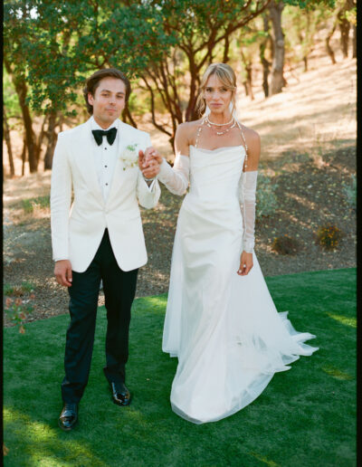 A couple dressed in formal wedding attire stands outdoors on grass, holding hands. The groom wears a white tuxedo, and the bride wears a white gown with long gloves.