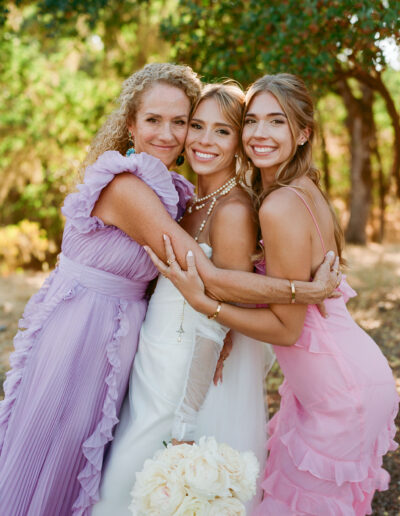 Three women in pastel dresses embrace outdoors, smiling at the camera. The woman in the center holds a bouquet of white flowers.