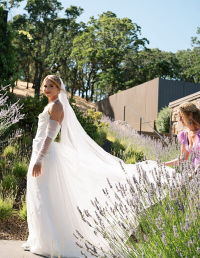 A bride in a white dress and veil stands on a garden path, turning back towards another woman adjusting her train. Lavender plants line the path, with trees and a building in the background.