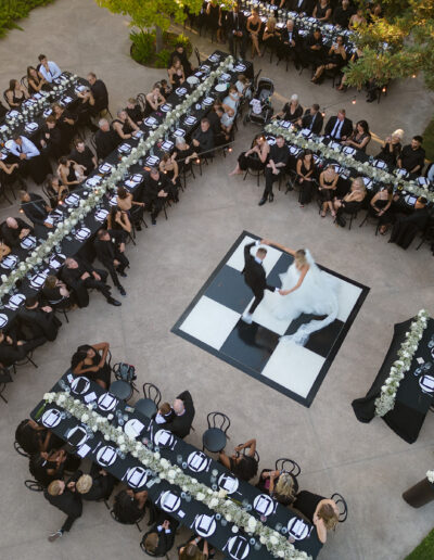 A bride and groom dance on a square checkered floor surrounded by guests seated at long tables in an outdoor venue.