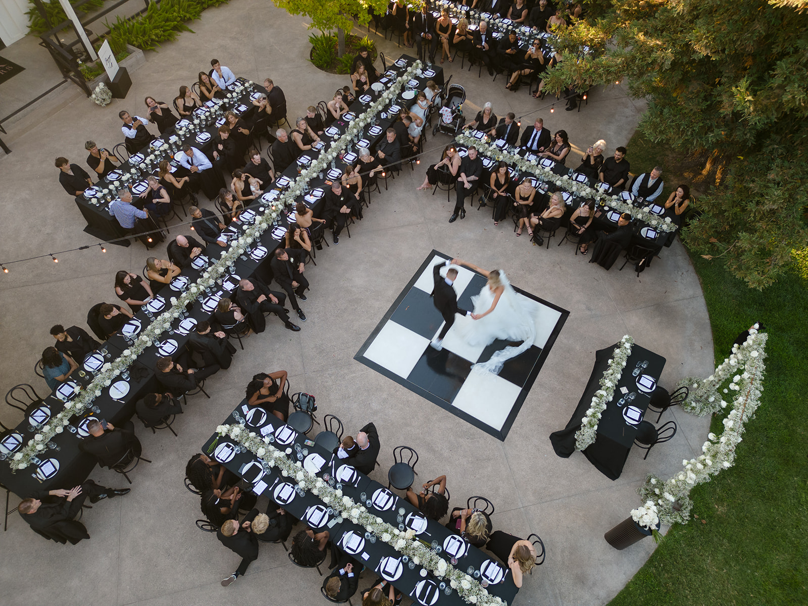 A bride and groom dance on a square checkered floor surrounded by guests seated at long tables in an outdoor venue.