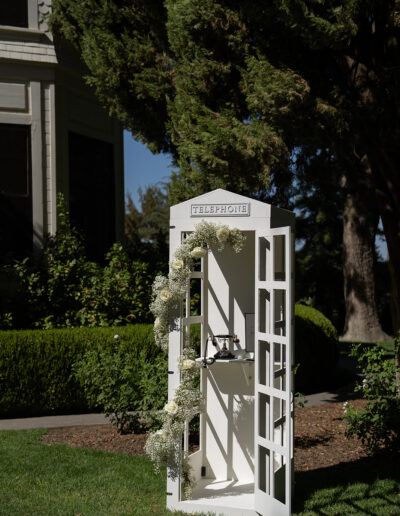 White decorative telephone booth adorned with flowers stands on a grass lawn near trees and a building.