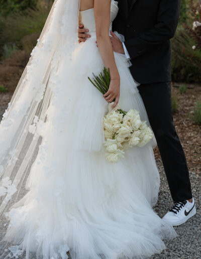 Bride and groom embrace outdoors; bride in white gown holds white roses, groom in black suit with sneakers.