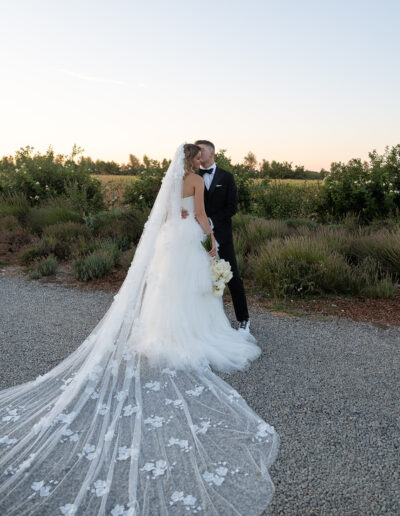 A bride and groom kiss outdoors, surrounded by greenery. The bride wears a long, lacy veil.