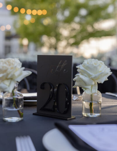 Outdoor table setting with a "Table 20" sign between two white roses in glass vases. Background features blurred trees and string lights.