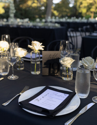 Elegantly set table with black tablecloth, menus, white roses in glass vases, and glassware at an outdoor event.