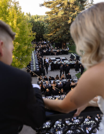 A couple observes a large outdoor wedding reception from a balcony. Long tables are surrounded by guests, with trees in the background.